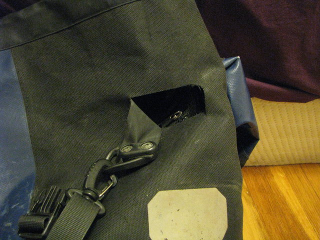 Close photo of one side of a blue and black bicycle pannier. A strip of fabric including the carrying strap's anchor has ripped partially away to the left, leaving a semi-rectangular hole with a view straight into the bag. The hole is about 4 cm high and 8 cm wide. The upper edge of the hole stretches farther to the left than the lower edge does.